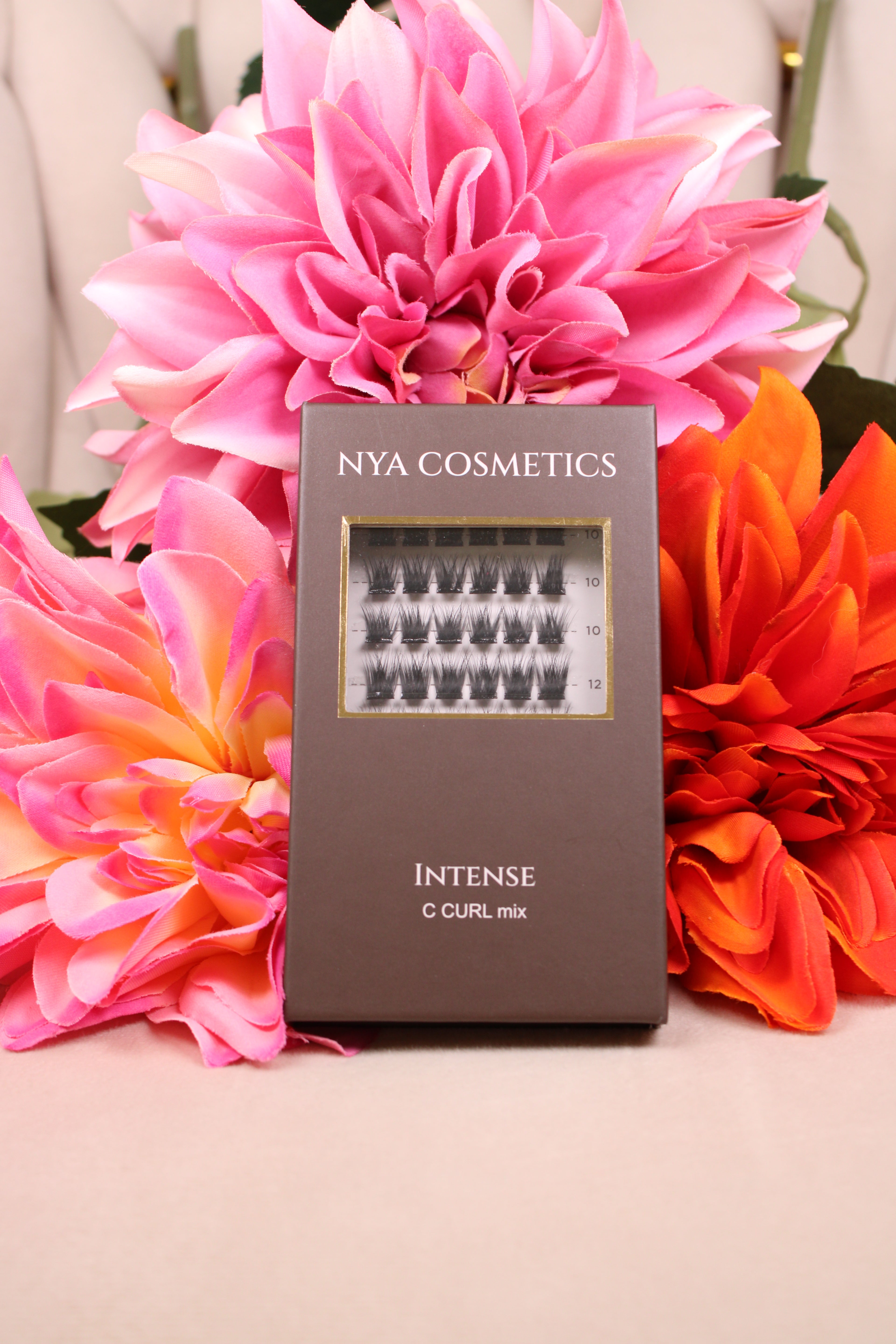 Boîte de faux cils auto-adhésifs NYA Cosmetics gamme Intense C Curl mix nouvelle collection posée devant des dahlias roses et oranges