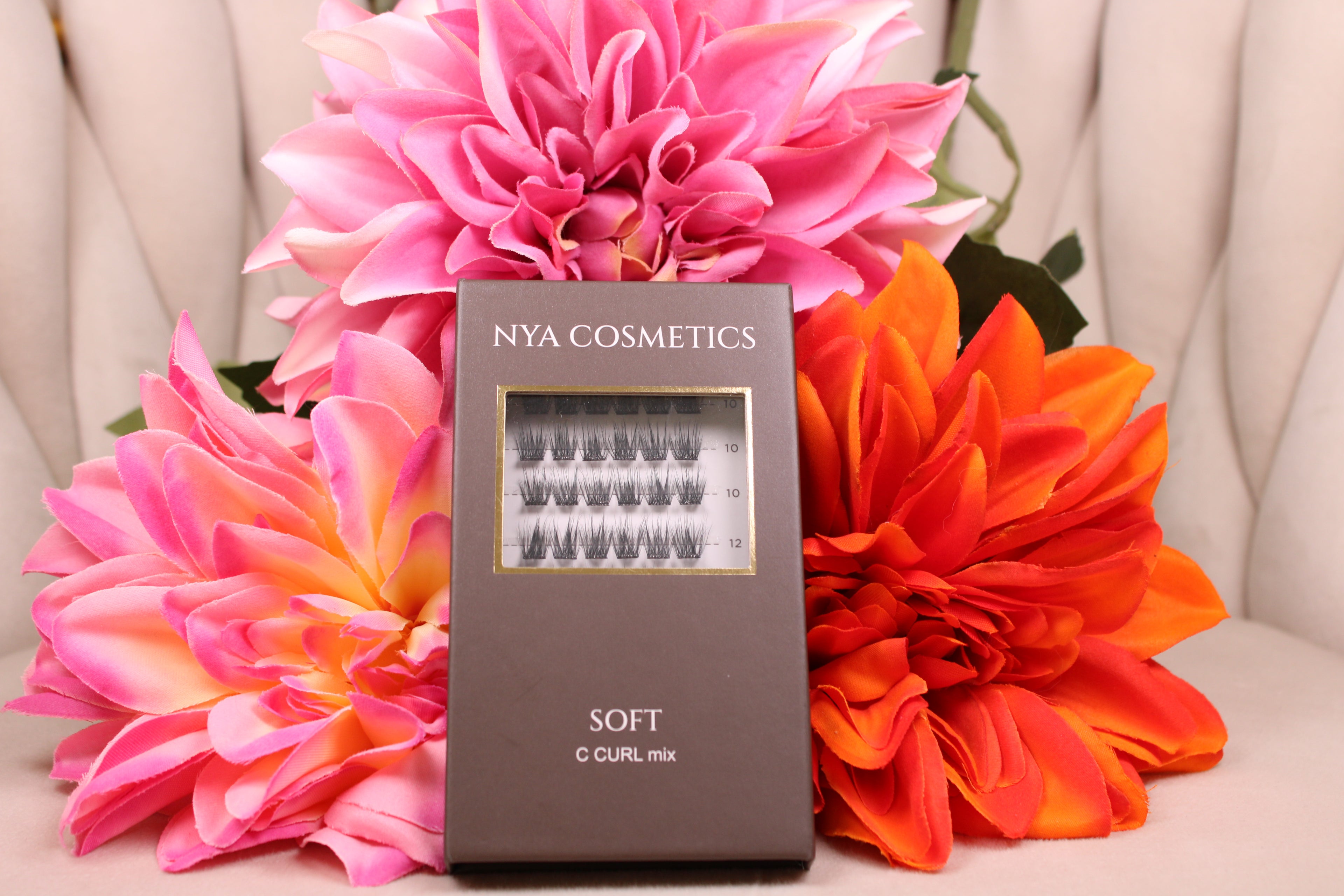 Boîte de faux cils auto-adhésifs NYA Cosmetics gamme Soft C Curl mix nouvelle collection posée devant des dahlias roses et oranges