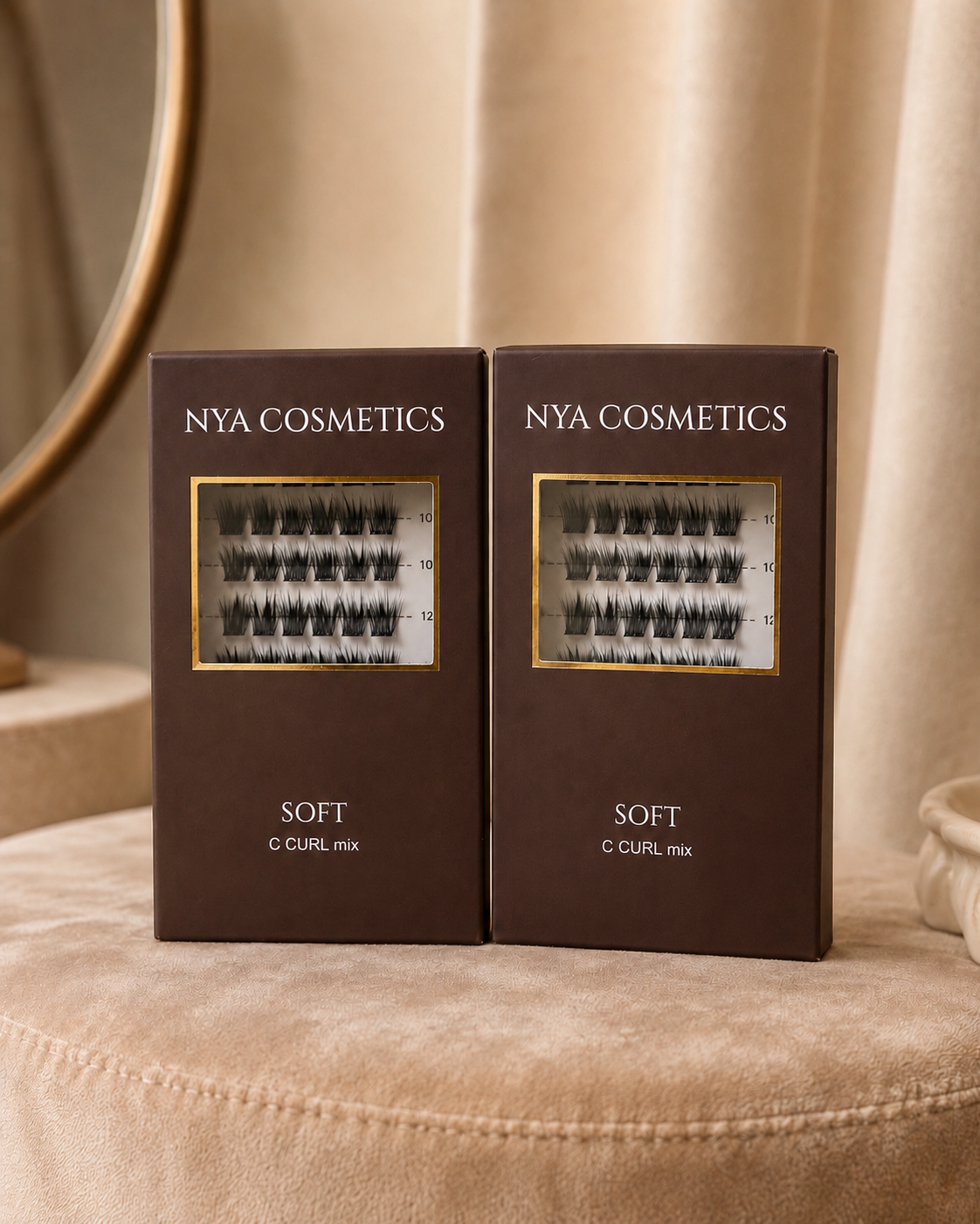 Duo de deux boîtes de faux cils auto-adhésifs NYA Cosmetics gamme Soft C Curl mix posées côte à côte sur un pouf velours beige avec miroir doré en arrière-plan nouvelle collection 2026